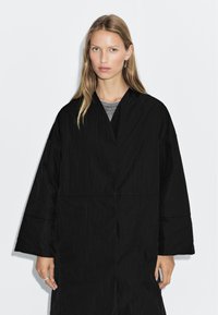 Black, oversized, textured coat with a V-neckline and wide sleeves. Worn over a grey shirt. The fabric appears crinkled, providing a casual look.
