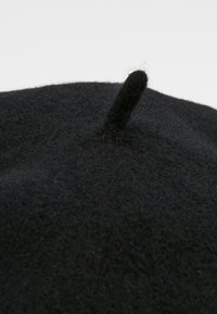 Beret preto de feltro com um pequeno e proeminente nó no topo. Apresenta uma textura suave e forma redonda, projetado para uso na cabeça.