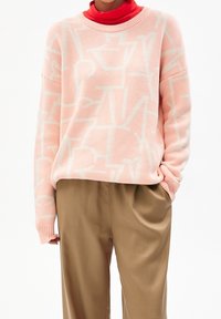 Persona che indossa un maglione rosa pastello con motivi astratti, una dolcevita rossa sotto e pantaloni beige larghi con le mani parzialmente nelle tasche.
