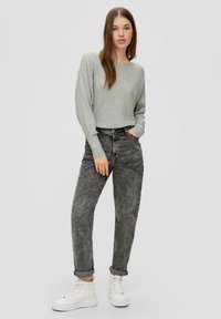 Hellgrauer gerippter Pullover, hochtaillierte dunkelgraue Jeans und weiße High-Top Sneaker. Model posiert mit einer Hand in der Tasche, lässiger Look.