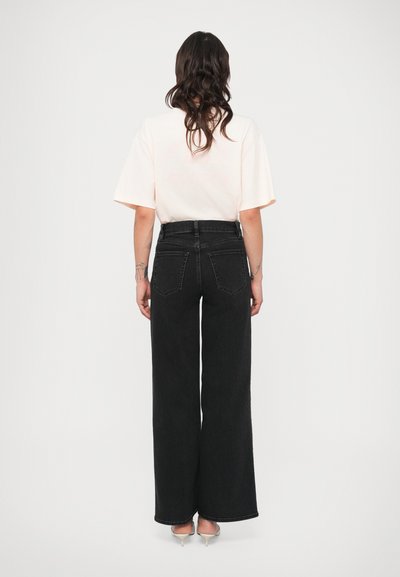 Jeans pretos de perna larga com cintura alta, com dois bolsos traseiros e textura lisa. Combinados com uma t-shirt de manga curta cor de rosa claro.