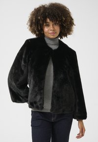 Melna mākslīgās kažokādas jaka ar noapaļotu kakla izgriezumu, īsām piedurknēm un mīkstu tekstūru. Nēsāta virs pelēkā turtlenecka, redzami tumši šorti.