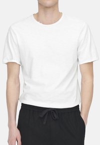 Weißes Baumwoll-T-Shirt mit kurzen Ärmeln und rundem Ausschnitt, mit einem leicht rohen Saum. Getragen mit schwarzen Jogginghosen mit Kordelzug.