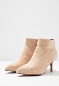 Botas de tobillo de ante beige con puntera afilada, tacón delgado y cierre de cremallera en la parte trasera. Textura suave, diseño minimalista.