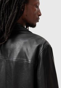 Schwarze Lederjacke mit glatter Textur, ausgestattet mit einem breiten Kragen und gesteppten Details auf dem Rücken. Die tiefen Farben verstärken ihr Aussehen.