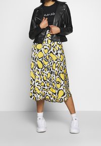 Černá kožená biker bunda přes černé tričko, doplněná žlutou a černou vzorovanou midi sukní a bílými teniskami.