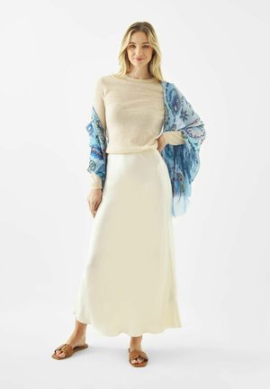 Vrouw staand, gekleed in een beige trui, lange crèmekleurige rok, blauw patroonachtige sjaal en bruine sandalen, tegen een witte achtergrond.