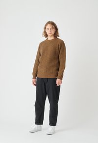 Brauner Strickpullover mit strukturiertem Muster, Rundhalsausschnitt, kombiniert mit schwarzen Hosen und weißen Sneakers, vor einem hellen Hintergrund stehend.