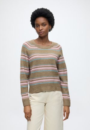 Femme aux cheveux courts et bouclés portant un pull beige avec des rayures horizontales roses et bleues et un pantalon couleur crème, debout devant un fond blanc.