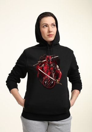 Person iført en sort hættetrøje med Deadpool-grafik, der holder to sværd, stående med hænderne i lommerne og kigger let til siden.