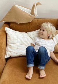 En tanfarvet plys snegl pude på en brun sofa, med et barn iført en hvid skjorte og blå jeans, der sidder tæt ved på beige puder.
