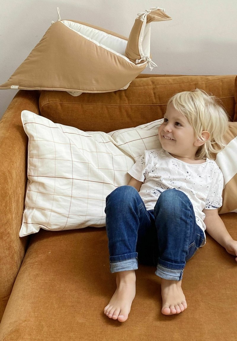 En tanfarvet plys snegl pude på en brun sofa, med et barn iført en hvid skjorte og blå jeans, der sidder tæt ved på beige puder.