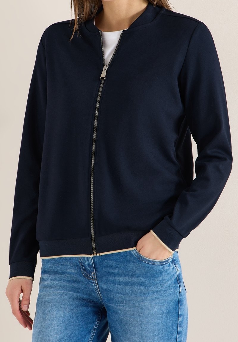 Femme portant une veste bleu marine zippée avec un bord beige sur une chemise blanche et un jean bleu, une main dans la poche.