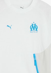 Balta sporta krekls ar ziliem akcentiem, ar Puma logo un Marseļas emblēmu, kā arī ar rāvējslēdzēja kabatu priekšpusē.