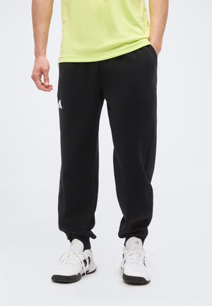 Personne portant un pantalon de jogging Adidas noir avec des poignets élastiques, un t-shirt vert clair et des baskets blanches avec des détails noirs, debout avec une main dans la poche.
