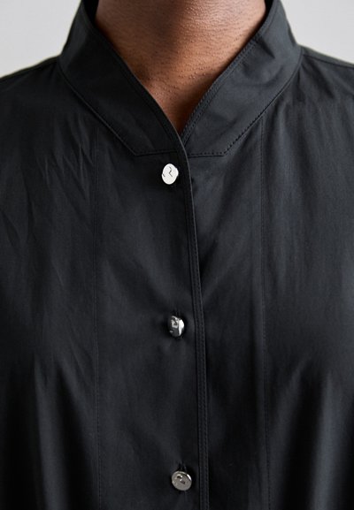 Camisa negra de botones con cuello alzado. Fabricada en un tejido suave con botones plateados y detalles de costura sutiles.
