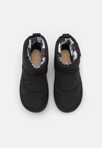 Paire de chaussons d'hiver noirs isolants à enfiler, avec extérieur rembourré et doublure en polaire beige douce, vus de dessus sur un fond blanc.