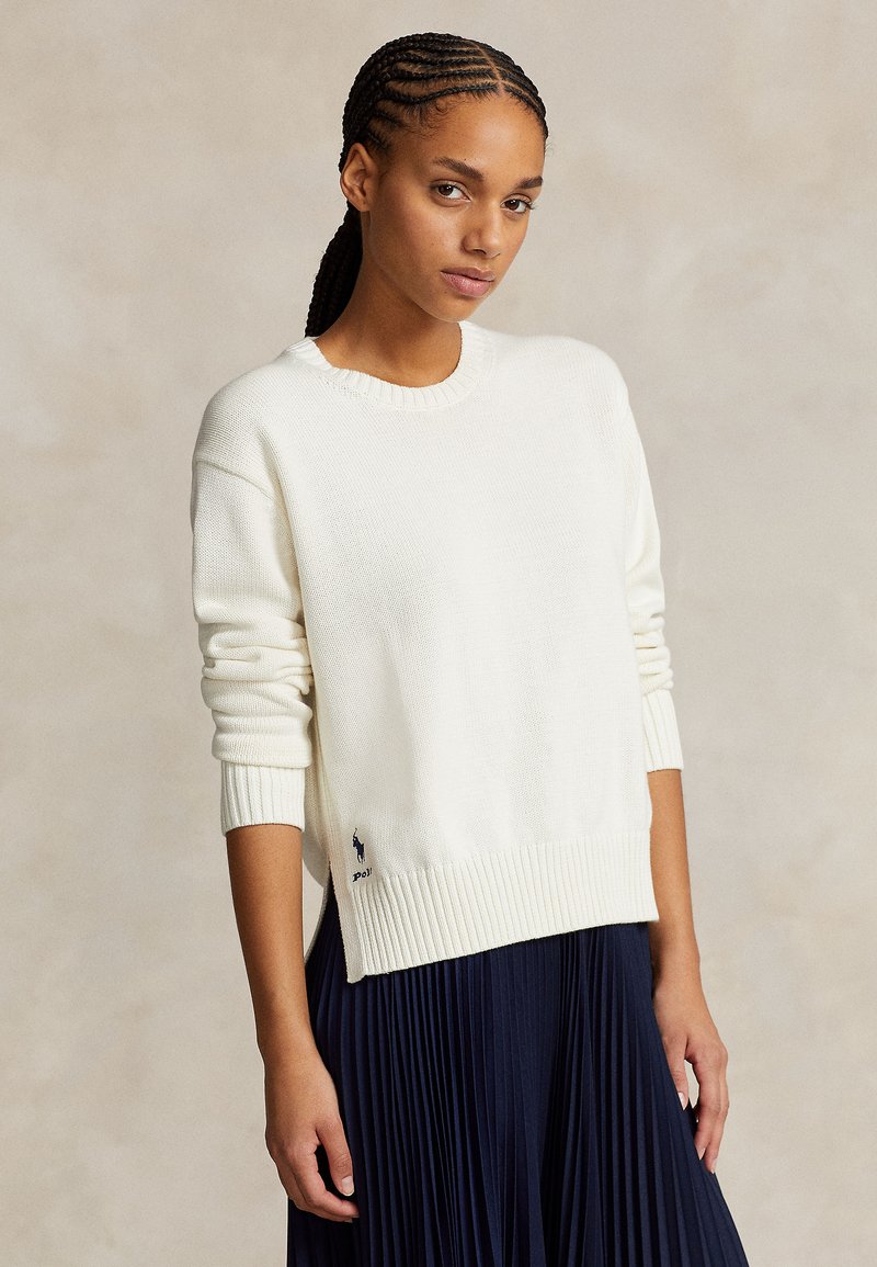 Balta, ribuotas megztinis su apvalia iškirpte, ilgomis rankovėmis. Apačia šiek tiek ilgesnė, šone yra mažas logotipo detalė.