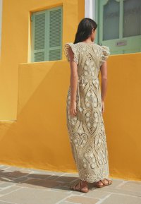 Robe maxi en dentelle beige avec des motifs floraux et géométriques, dotée de manches volantes et d'une taille cintrée, sur fond de mur jaune.