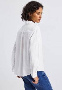 Femme avec des cheveux bouclés courts, portant une chemise blanche boutonnée ample et un jean bleu, se tenant avec le dos partiellement tourné.
