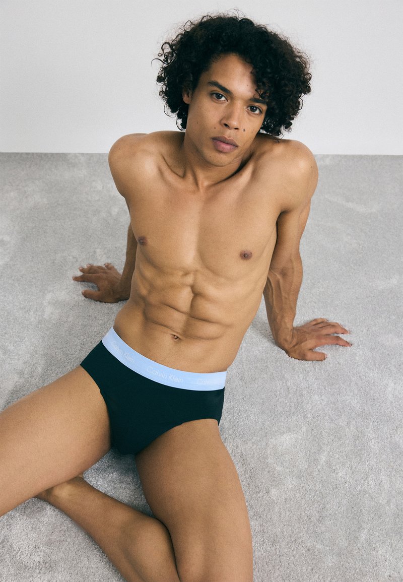 Fekete pamut bugyi világoskék derékrésszel, amelyen a "Calvin Klein" logó szerepel. A modell egy világosszürke felületen ül, kidolgozott fizikai alakkal.