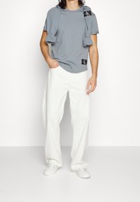 Hombre con camiseta gris y suéter sobre los hombros, pantalones blancos holgados y zapatillas blancas, de pie con una mano en el bolsillo.