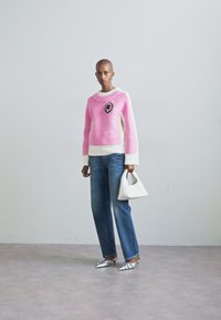 Pull en tricot rose avec texture côtelée et patch floral noir, associé à un jean bleu clair, un sac à main blanc et des chaussures à bouts pointus argentées.