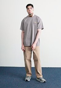 Grey cotton t-shirt with short sleeves, featuring a small logo. Paired with beige trousers and gray sneakers on a blue carpet background.