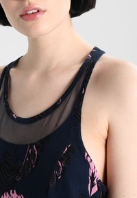 Granatowy bezrękawnik z siateczkowym dekoltem, zdobiony różowymi kwiatowymi wzorami i luźnym krojem. Faktura wydaje się gładka z subtelnymi detalami.