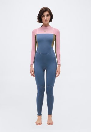 Γυναίκα που στέκεται ξυπόλητη φορώντας ολόσωμη στολή κατάδυσης με ροζ μανίκια, μπλε κορμό και πόδια, και ελιά πλαϊνά πάνελ σε λευκό φόντο.