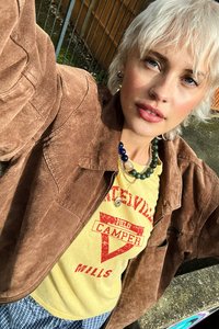 Brown suede jacket, yellow graphic tee with red design, layered necklaces with multicolored beads, plaid blue and white bottom.