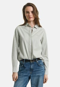 Femme aux longs cheveux bruns portant une chemise boutonnée blanche et grise à rayures verticales rentrée dans un jean bleu avec une ceinture noire.