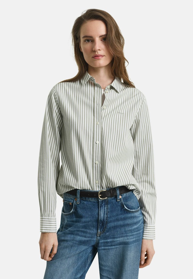 Femme aux longs cheveux bruns portant une chemise boutonnée blanche et grise à rayures verticales rentrée dans un jean bleu avec une ceinture noire.