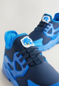 Blå sportskor med marinblått tyg, texturerade gummidetaljer, en tecknad karaktärs emblem och kontrasterande blå detaljer på sulan.