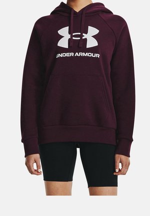 Osoba w bordowej bluzie z kapturem Under Armour z białym logo i napisem, w czarnych spodenkach kolarskich, stojąca na jednolitym tle.