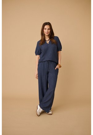 Pikkade pruunide juustega naine, kes kannab mereväe sinist tekstuuriga kahetükki komplekti, valgeid tossusid, kaelakeed ja pruuni käevõru beeži tausta ees.