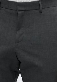 Gros plan sur un pantalon formel gris foncé porté avec une chemise blanche, montrant la taille, la boucle de ceinture et le pli avant.
