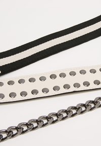 Tre cinturini distinti: un cinturino in tessuto nero con strisce bianche, un cinturino in eco-pelle bianco con borchie metalliche e un cinturino a catena nera lucida.