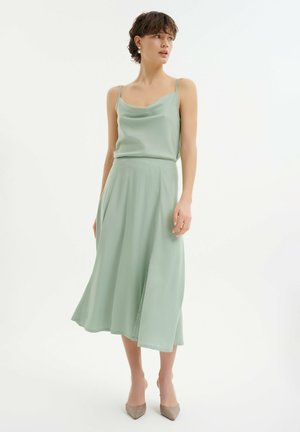 Cowl neck dress in light green fabric with a flowing silhouette, featuring thin straps and a knee-length hemline that opens at one side.
