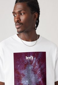 Mand iført hvid Levi's t-shirt med et grafisk motiv af en lilla skov og en stjernehimmel, som ser til venstre mod en ensfarvet baggrund.