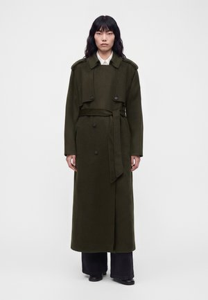 Femme aux longs cheveux foncés portant un long trench-coat ceinturé vert foncé sur un pantalon noir et des chaussures noires, debout devant un fond blanc uni.