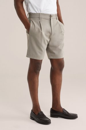 Mees kannab beežikaid voltidega lühikesi pükse küljetaskutega, valget sisse toppinud särki ja musti paelu, seisab neutraalsel taustal.