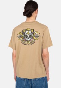 Tan cotton t-shirt with a graphic design featuring a skull and snakes on the back. Short sleeves and a relaxed fit.