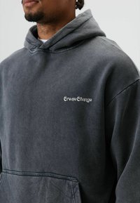 Sweat à capuche gris en tissu doux, avec une poche avant, une capuche avec cordon de serrage et un logo brodé "Create Change" sur la poitrine.