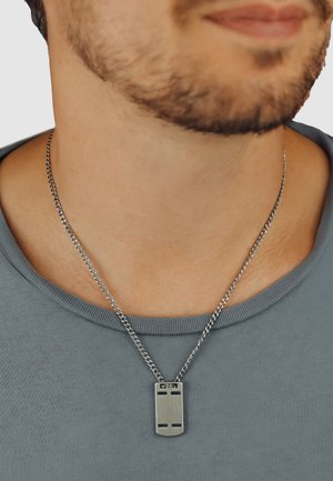 Silberne Metalkette mit einem rechteckigen Anhänger, der zwei ausgeschnittene Designs aufweist, getragen über einem grauen Shirt. Der Anhänger hat eine gebürstete Oberfläche.