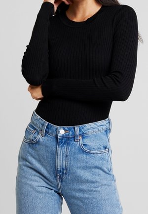 Femme portant un haut noir moulant à manches longues côtelé, rentré dans un jean bleu clair taille haute, bras croisés à la taille.