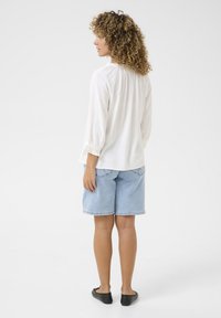 Blusa bianca a maniche lunghe con dettaglio arricciato sul retro, abbinata a pantaloni shorts in denim chiaro e scarpe slip-on nere, vista da dietro.