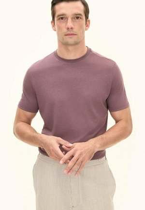 Mężczyzna ubrany w fioletową koszulkę z krótkim rękawem i jasnobeżowe spodnie w paski, stoi z rękami złożonymi przed sobą na jednolitym tle.