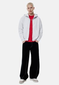 Felpa grigia con zip, t-shirt rossa, pantaloni larghi neri con cuciture bianche e sneakers beige. Tessuti lisci e vestibilità casual.