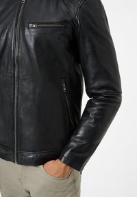 Schwarze Lederjacke mit tailliertem Design, zwei Reißverschlusstaschen, glatter Textur und sichtbaren Nähten entlang der Kanten.